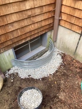 A metal vent well with gravel around a low foundation opening beside a downspout