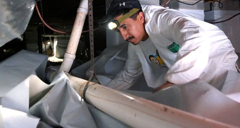 A technician wearing a headlamp fitting white liner material inside a crawl space
