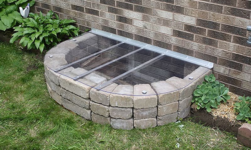 An existing covered window well beside a brick foundation