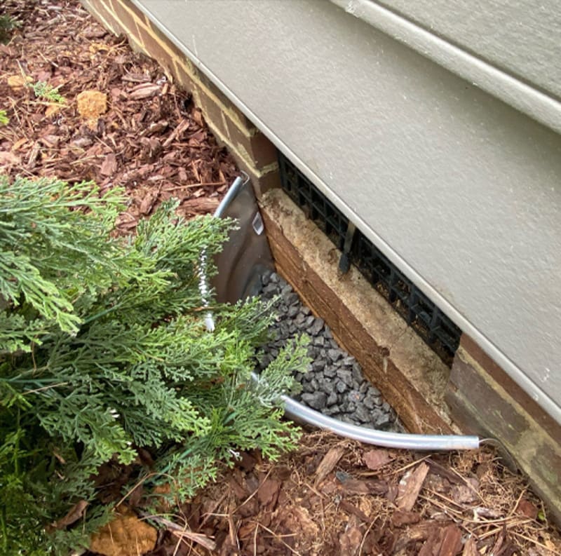 A metal vent well with gravel around a low foundation vent beside landscaping