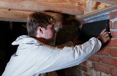 A technician fitting a dark panel into a crawl space vent opening