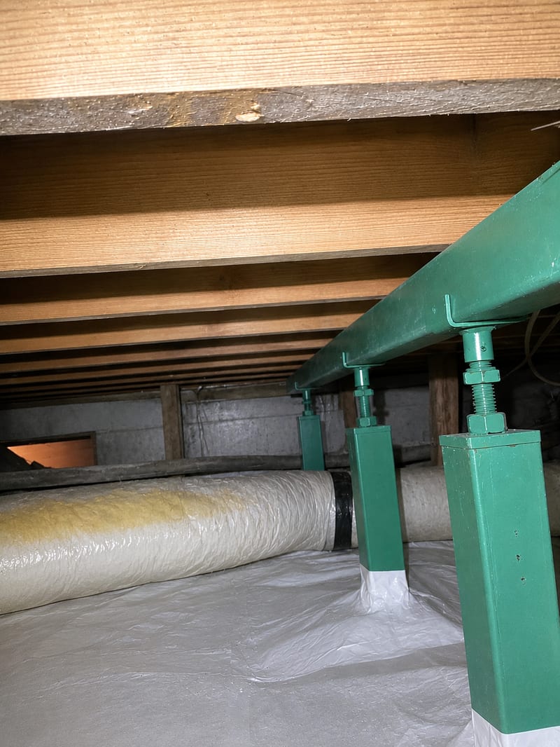 A galvanized steel crawl space jack supporting a wooden floor beam above a bare dirt floor